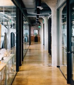 A modern office hallway with concrete pillars and glass walls, looking towards the exit.