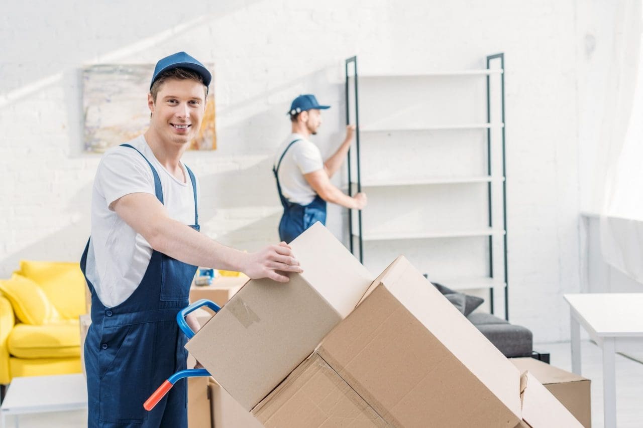 moving company two movers in uniform transporting cardboard boxes and furniture in apartment e1670221445352